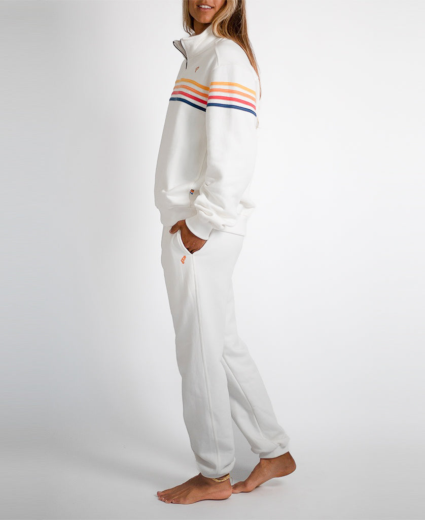 A person stands barefoot in the Golden Breed Long Island 1/4 Zip in Off White—cotton fleece with orange, yellow, and blue stripes and the Retro Golden Breed Logo—hands in pockets, facing sideways against a plain white background.