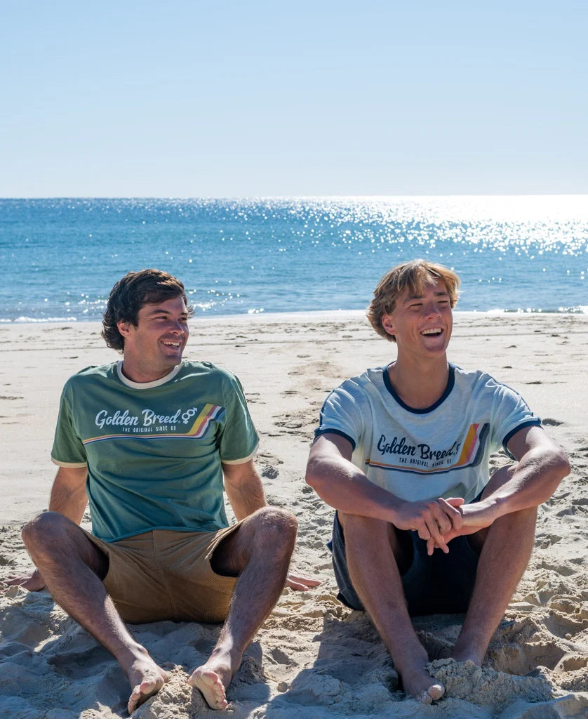 Two men sitting on a sandy beach smiling and relaxing by the ocean wearing casual surfwear, one wearing a Retro Ringer Tee | Ice with navy blue accents under clear blue skies.