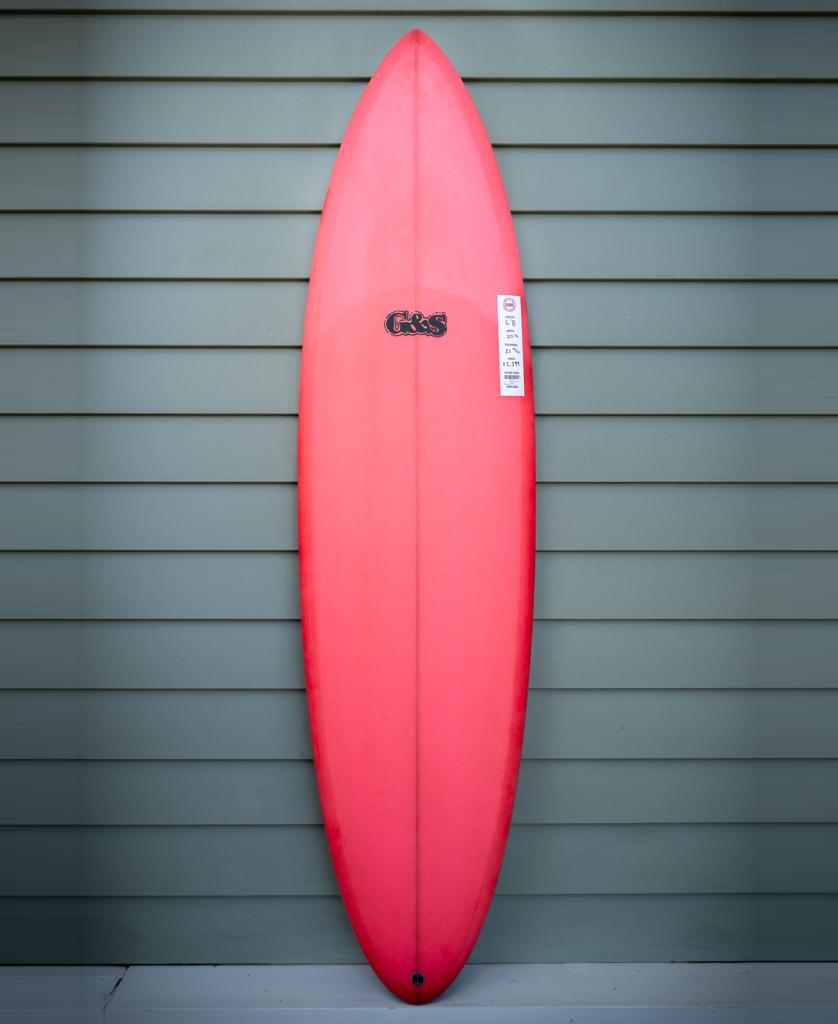The G&S 68 Cool Tool Mid Twin | Red by Gordon & Smith, featuring a black logo and white label, stands upright against a light green horizontal siding wall—an iconic surfboard from the 70s.