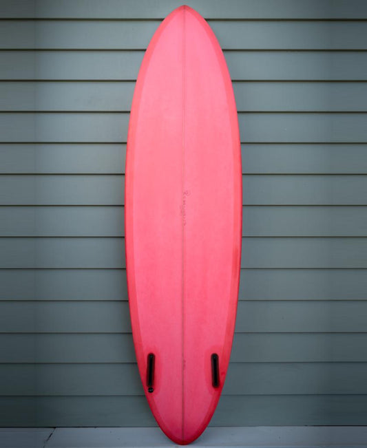The Gordon & Smith G&S 68 Cool Tool Mid Twin surfboard, in red and featuring two black fins, stands upright against a wall with horizontal green siding.