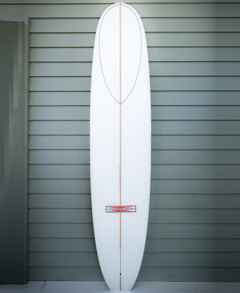 The Gordon & Smith G&S 96 Perfecto Polish Finish | White longboard, featuring a brown stringer, stands vertically against a gray wooden wall, designed for smooth nose riding.
