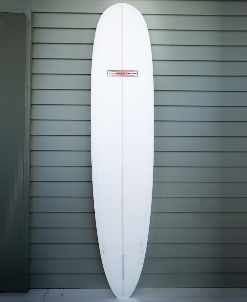 The Gordon & Smith G&S 94 Nosemaster Polish Finish in blue and white stands upright against a light green paneled wall, ideal for nose riding on mellow waves.