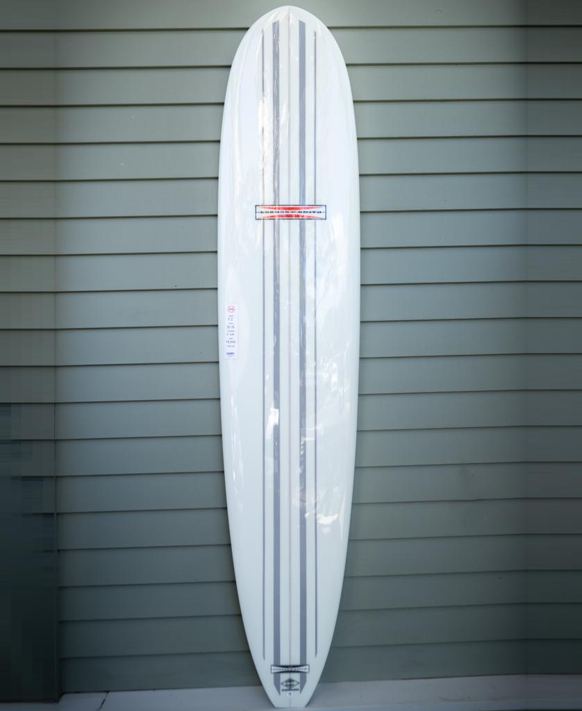 The Gordon & Smith G&S 91 Nosemaster Polish Finish | White/Grey longboard, featuring three grey stripes, stands upright against a light green wall—ideal for classic nose riding.