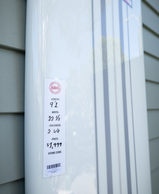 A G&S 91 Nosemaster Polish Finish longboard by Gordon & Smith in white/grey leans against a wall, price tag displaying $1,199, dimensions, and store code.