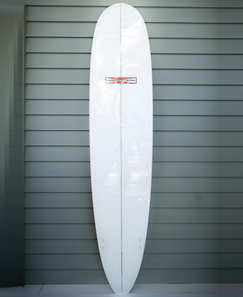 The G&S 91 Nosemaster Polish Finish longboard by Gordon & Smith, in white/grey with a red and black label, stands upright against a light gray paneled wall—ideal for classic nose riding.