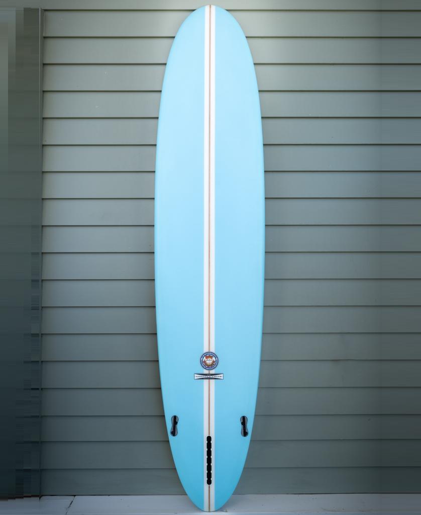 The Gordon & Smith G&S 91 Hotdogger Matte Finish | Blue longboard surfboard stands upright against a gray paneled wall, featuring a light blue color and a white center stripe.