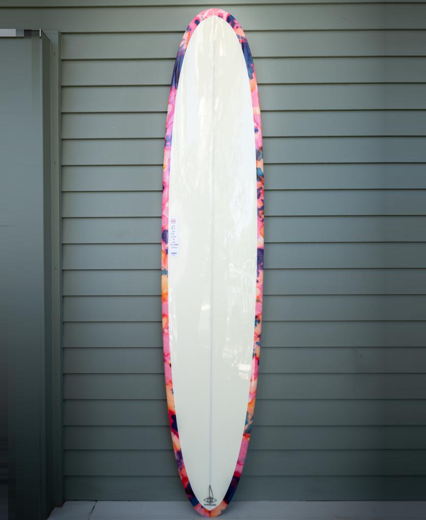 The Gordon & Smith G&S 93 Chick Board Polish Finish in Pink Tie Dye stands upright against a gray paneled wall, its vibrant pink, orange, and blue border ready to catch any size wave.