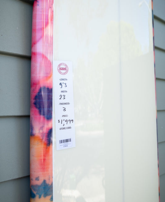 The Gordon & Smith G&S 93 Chick Board Polish Finish in Pink Tie Dye stands upright against a wall, with a tag showing its size (9’3” x 23”) and price ($1,999). Perfect for catching any size wave.