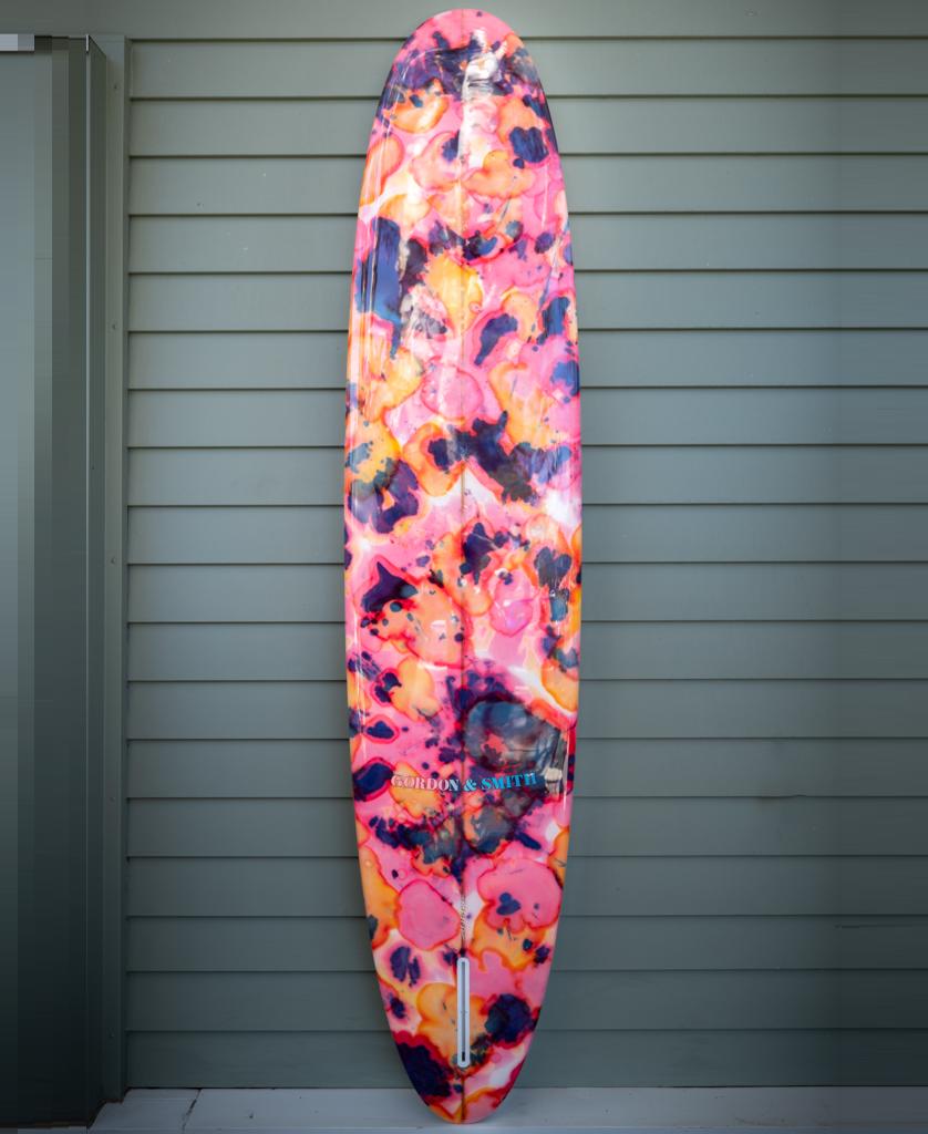 The Gordon & Smith G&S 93 Chick Board Polish Finish | Pink Tie Dye longboard, featuring a vibrant pink tie-dye pattern, leans against a light green paneled wall—ideal for catching waves of any size.