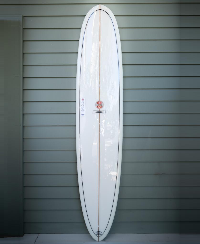 A G&S 93 Dane Wilson Polish Finish longboard by Gordon & Smith in white, featuring a thin wooden center stringer, stands upright against a light gray paneled wall.