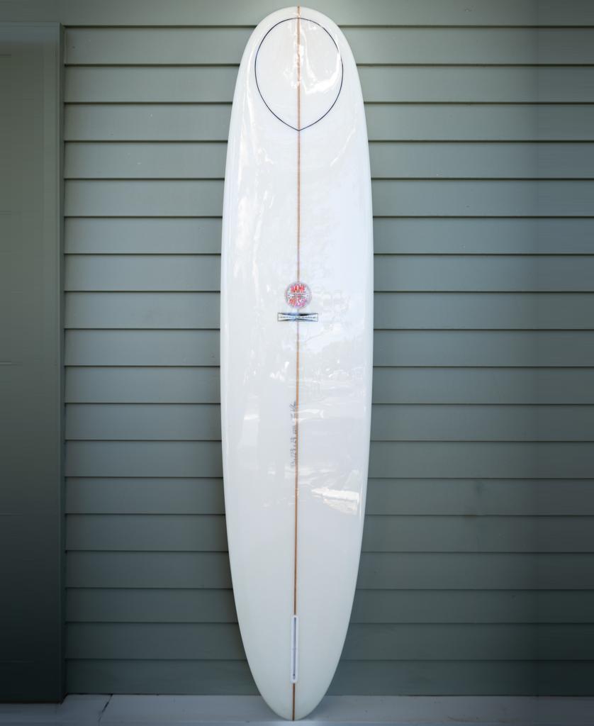 The Gordon & Smith G&S 93 Dane Wilson Polish Finish | White longboard, featuring a sleek white design and classic polish, stands upright against a light gray paneled wall, reflecting timeless surf style.