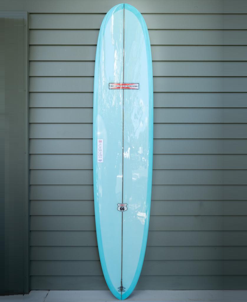 The Gordon & Smith G&S 91 Route 66 longboard surfboard with a polish finish stands upright against a gray wooden wall, featuring a single fin setup and leash plug in light blue.