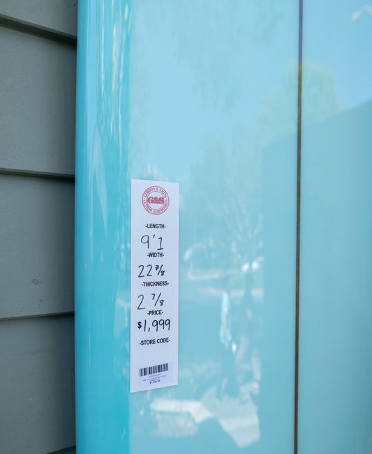The G&S 91 Route 66 by Gordon & Smith is a blue longboard surfboard with a white label showing its 9’1” length, 22 7/8” width, and 2 7/8” thickness, priced at $1,999. Ideal for riders needing extra paddle power.