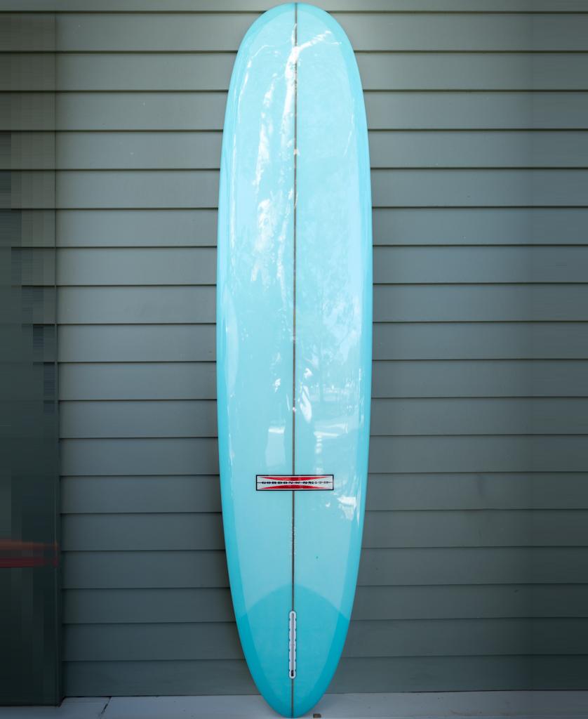 The Gordon & Smith G&S 91 Route 66 | Polish Finish longboard, featuring a light blue single fin design, stands upright against a wall with horizontal gray siding, ready for smooth rides and paddle power.