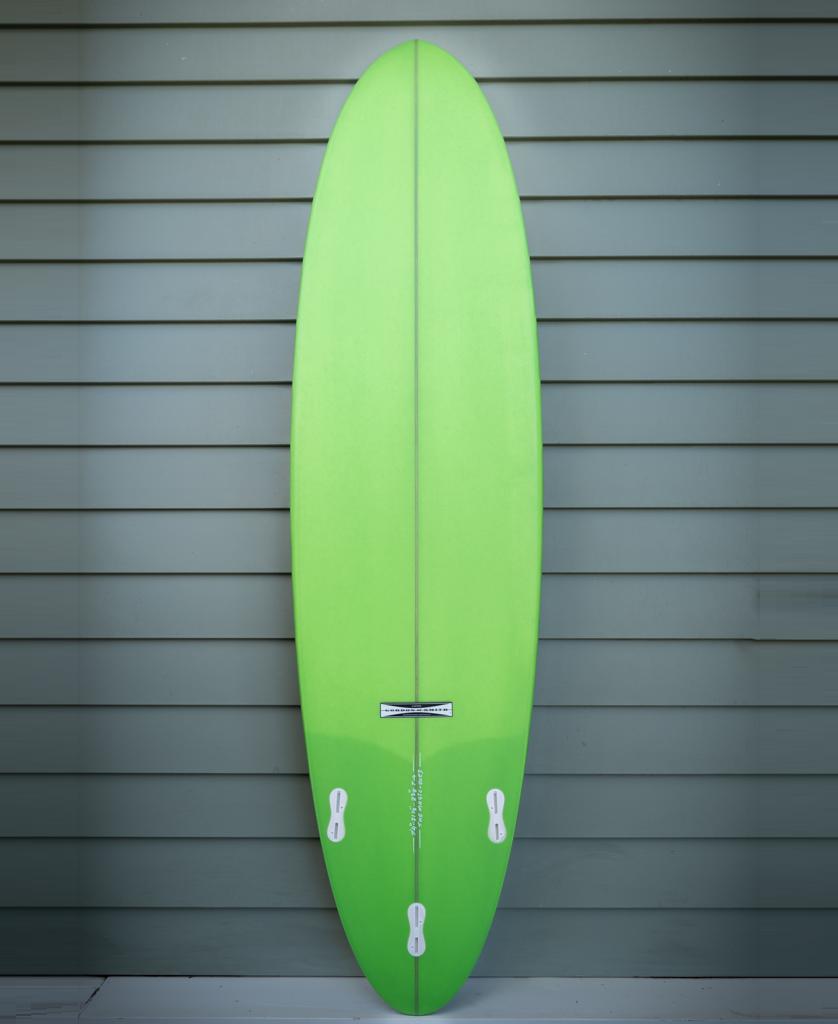 The Gordon & Smith G&S 74 Magic Polish Finish surfboard in white and lime stands upright with three white fins against gray horizontal siding.