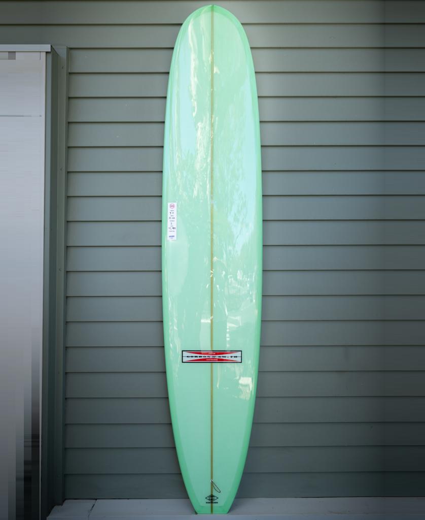 The Gordon & Smith G&S 98 Easy Rider Polish Finish longboard in lime, featuring a wooden stringer, stands upright against a gray paneled wall.