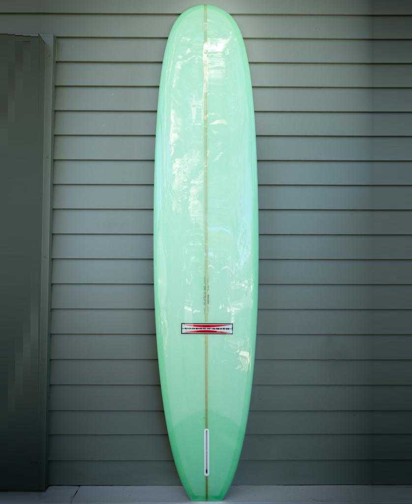 The Gordon & Smith G&S 98 Easy Rider Polish Finish in lime stands upright against a gray wood-paneled wall, showcasing its user-friendly design and single fin box with a nod to classic surf style.