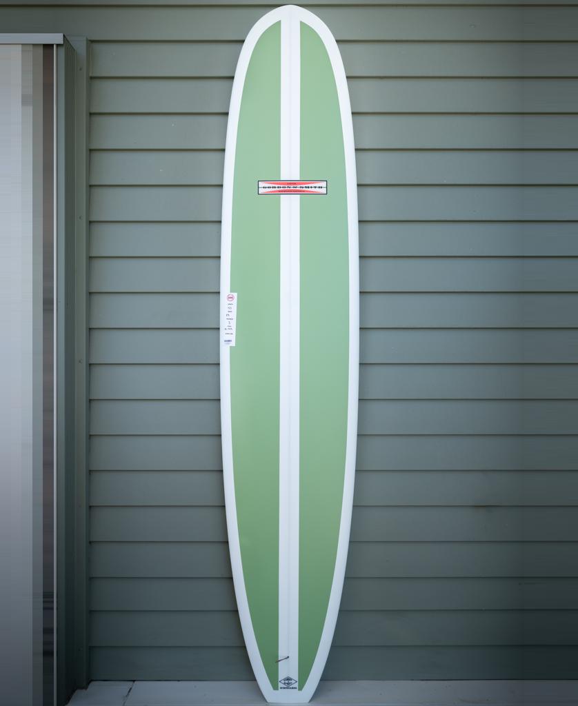 The Gordon & Smith G&S 93 Easy Rider Polish Finish longboard in green with white edges stands upright against a light gray paneled wall.