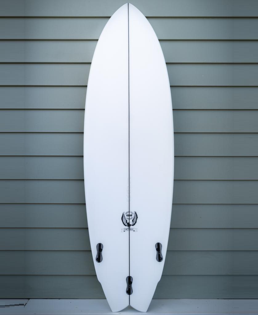 A Gordon & Smith G&S 64 Power Twin Matte Finish performance board in white with a fish tail and three black fins stands upright against a gray horizontal plank wall.