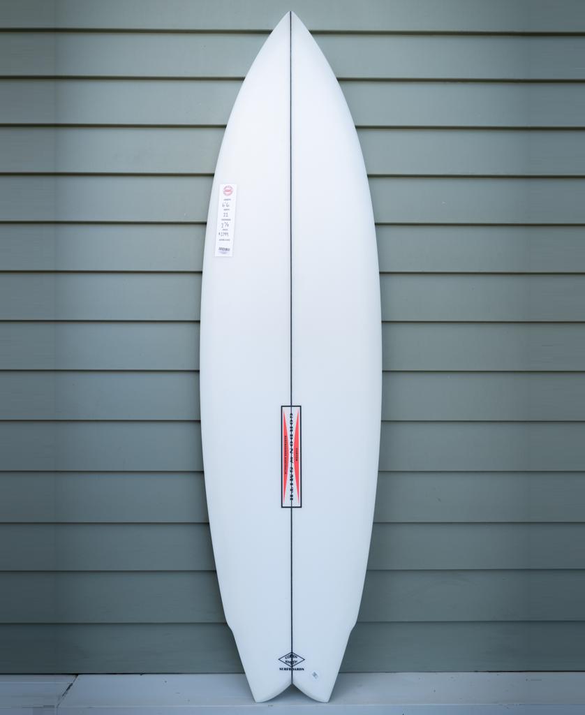 The Gordon & Smith G&S 66 Power Twin Matte Finish surfboard in white stands upright against a gray horizontal panel wall.