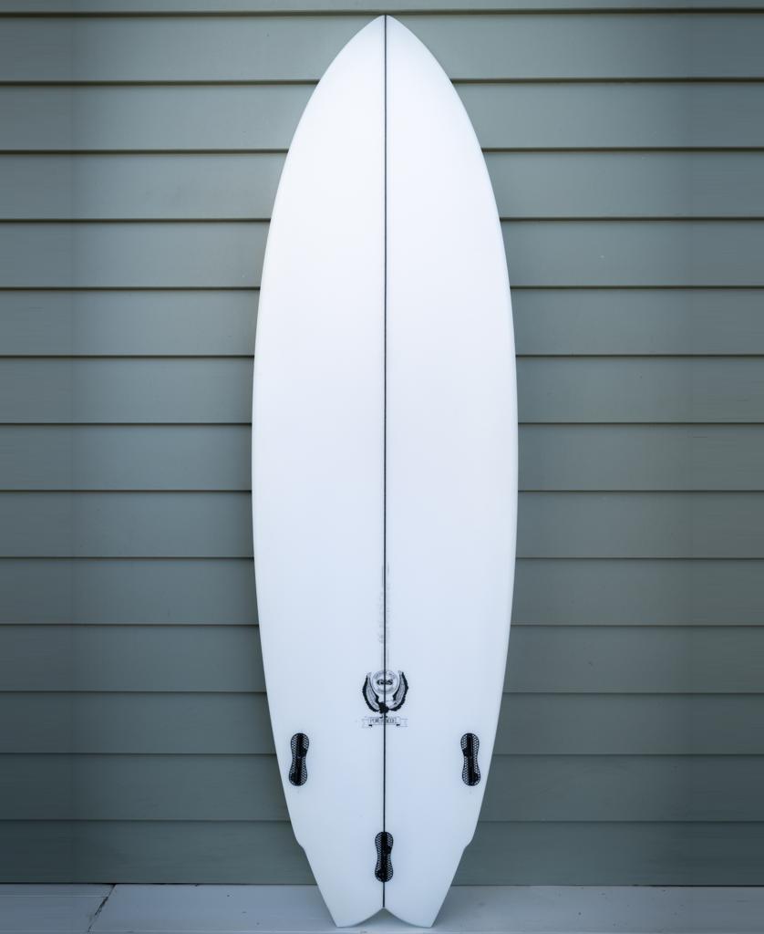 A Gordon & Smith G&S 66 Power Twin Matte Finish surfboard in white with black fins stands upright against a light green, horizontally paneled wall.