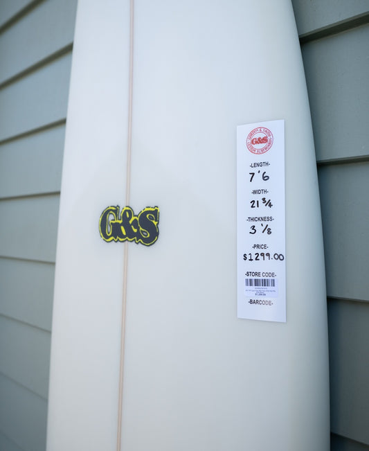 The G&S 76 Cool Tool Mid Twin in Off White by Gordon & Smith, priced at $1,291, leans against a gray paneled wall—perfect for surfers seeking superior wave-catching performance.