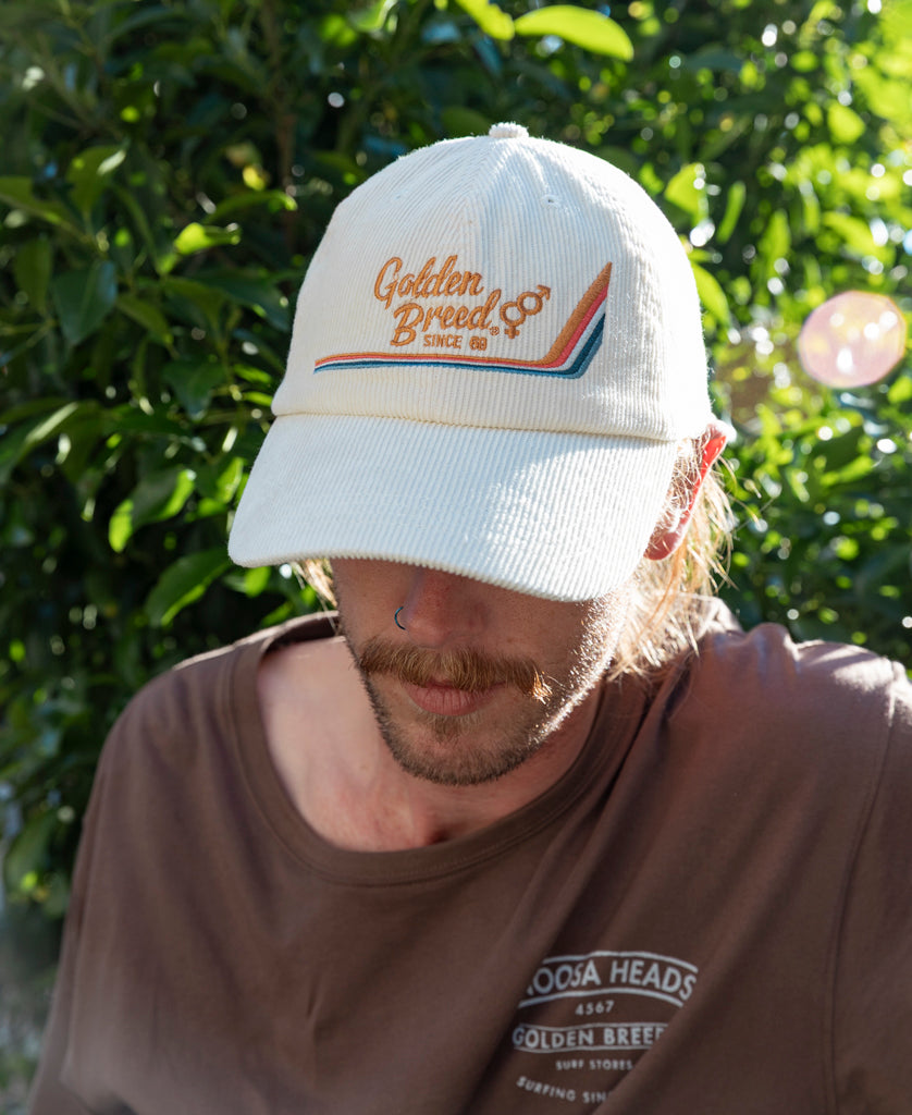 A mustached person wears a Golden Breed Hancock Cord Cap in off white and a brown Noosa Heads t-shirt, standing outdoors with green foliage in the background.