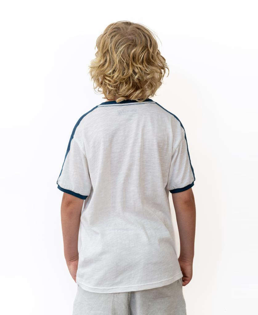 A young boy with curly blonde hair stands facing away, wearing the Golden Breed Flinders Rider Kids Tee in Off White and light-colored shorts, set against a plain white background.