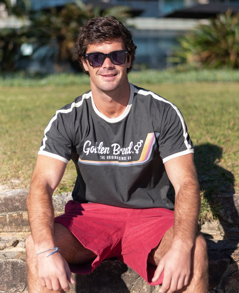 A man in sunglasses, a dark Retro Ringer Tee | Wash Black by Golden Breed, and red shorts sits on outdoor steps with grass and plants behind him, capturing laid-back 70s vibes.