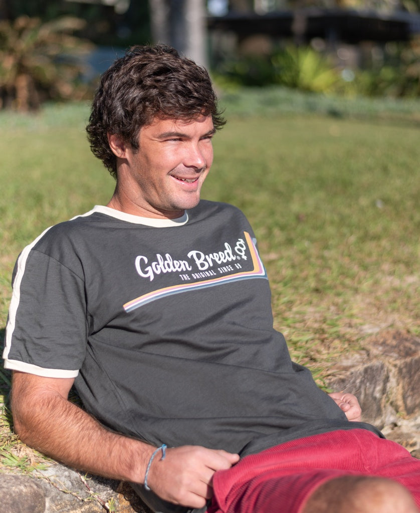 A man with brown hair wearing a Golden Breed Retro Ringer Tee in Wash Black and red shorts sits on a low stone wall outdoors in sunlight, giving off strong 70s vibes.