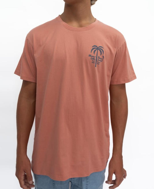 A person wearing the Golden Breed Manly Palm Loco Tee in Rose, a 100% cotton salmon T-shirt featuring a small palm tree and Miami Beach graphic on the left chest, pairs it with light blue jeans against a white background.