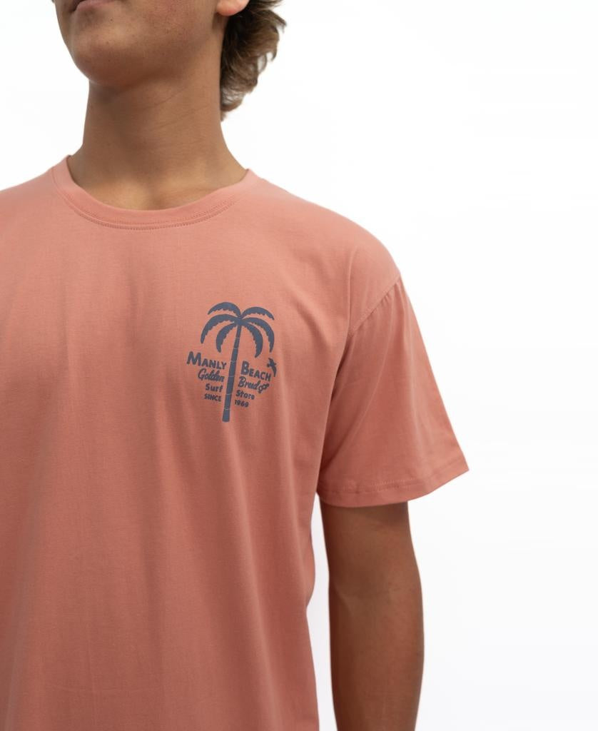 Someone wears the Golden Breed Manly Palm Loco Tee | Rose, a 100% cotton shirt in salmon pink with a blue palm tree and Manly Beach, Chill, Surf, Break, Since 1978 text on the left chest.