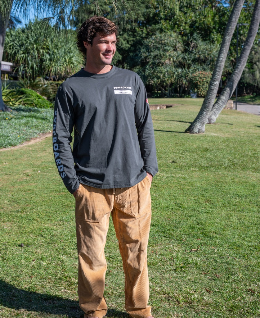 A man stands on grass wearing a long-sleeve dark shirt with text and Golden Breeds George Cord Pant in Dark Camel, featuring wide legs and a patch pocket, set against trees and a walkway.