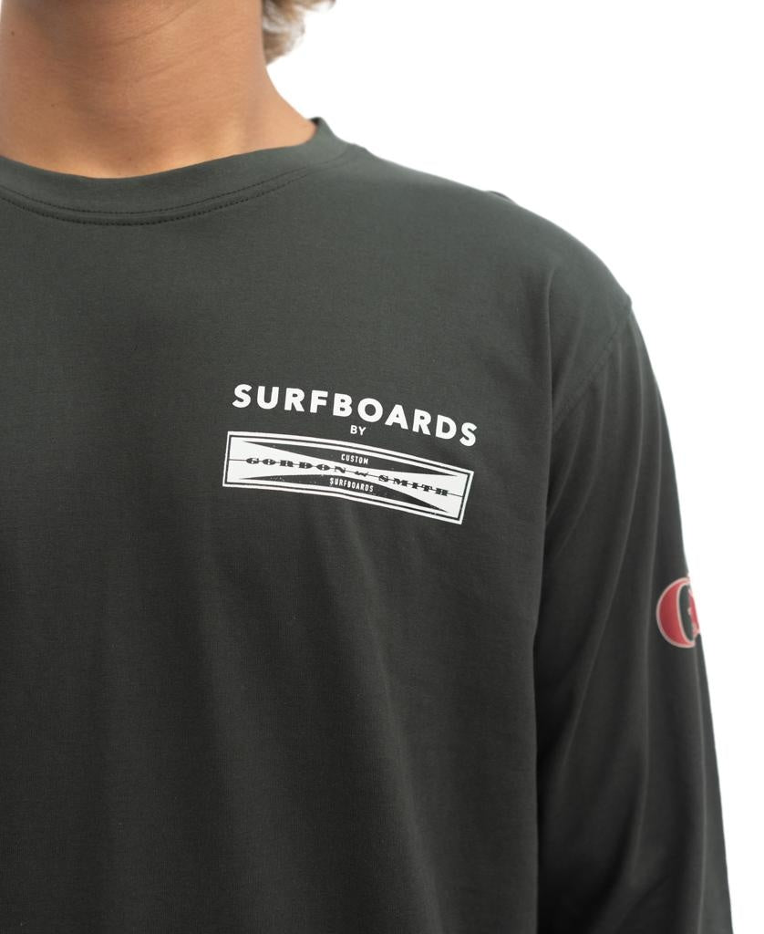 A person wears the G&S Surfboards Comp LS Tee in Wash Black by Gordon & Smith, featuring the SURFBOARDS text and logo on the chest, a Champion sleeve logo, and celebrates true surf heritage.