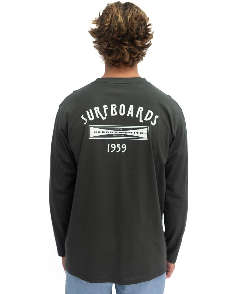Person with wavy hair wears the G&S Surfboards Comp LS Tee in Wash Black by Gordon & Smith, featuring SURFBOARDS 1959 on the back, celebrating surf heritage against a plain white background.