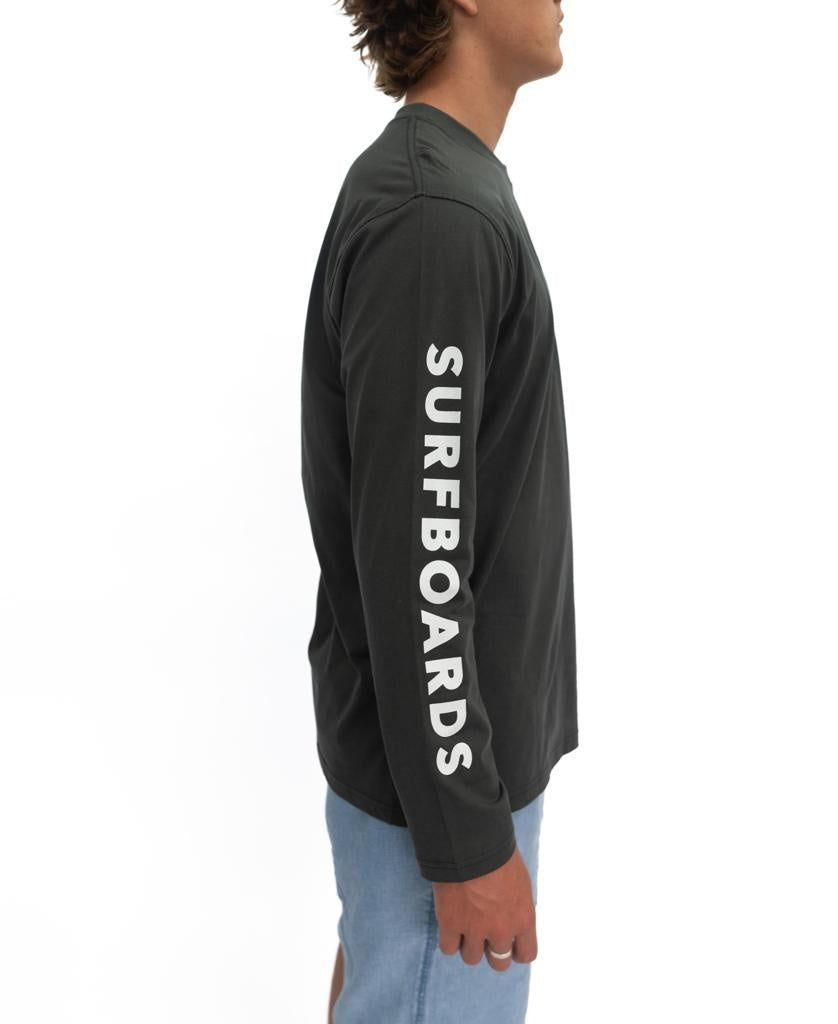 A person wears the Gordon & Smith G&S Surfboards Comp LS Tee in wash black, featuring SURFBOARDS in white on the right sleeve, paired with light blue shorts, standing against a white background.