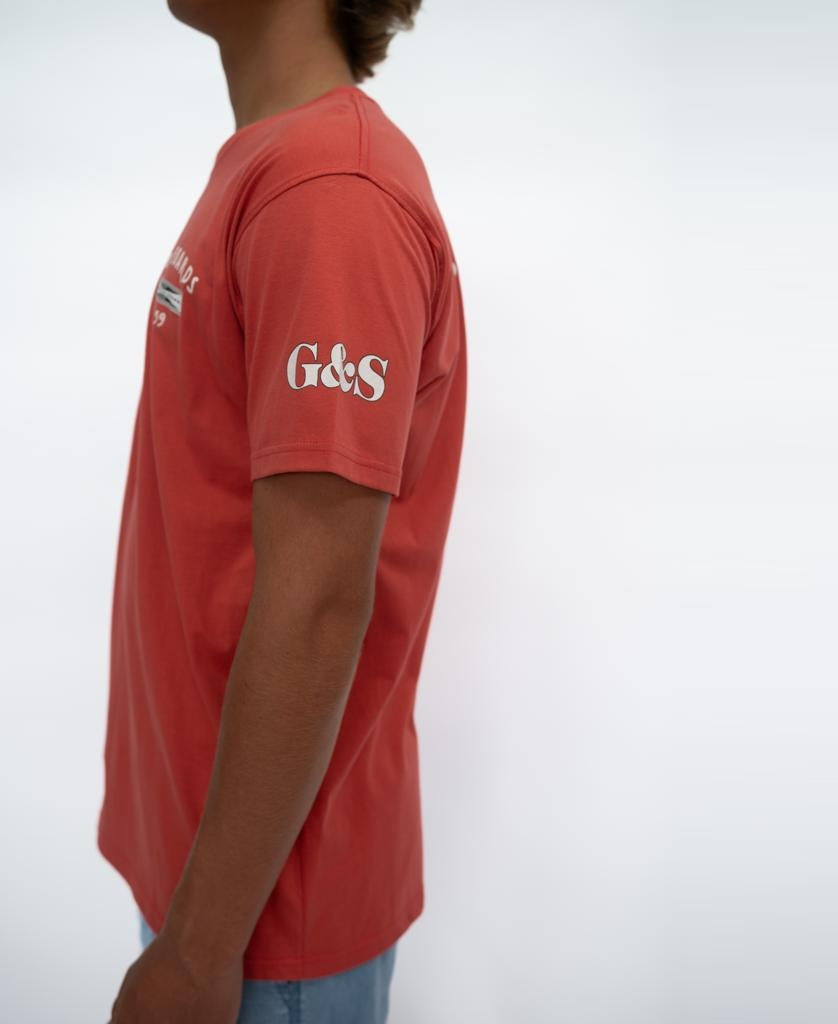 A person wearing a Wash Red G&S Surfboards Tee by Gordon & Smith with G&S on the left sleeve, standing against a plain white background.