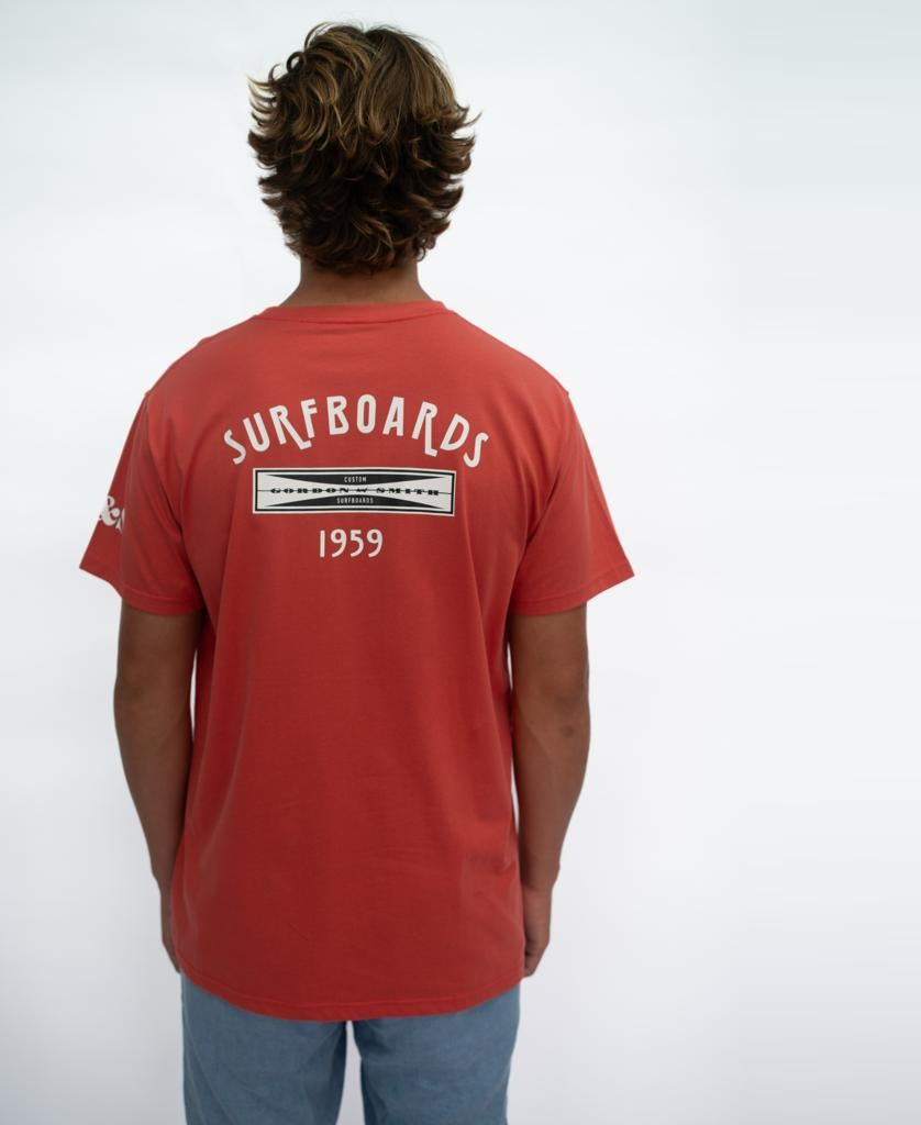 A person stands with their back to the camera, wearing a Gordon & Smith G&S Surfboards Tee in Wash Red with SURFBOARDS 1959 on the back, paired with light blue shorts against a plain white background.