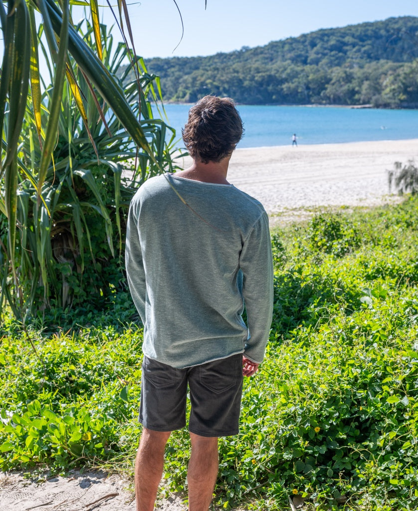Wearing a Golden Breed Classic Rag Top in Surf Green Marle and shorts, a person stands on grass gazing at the sandy beach and ocean with hills beyond, embodying an effortless surf style.