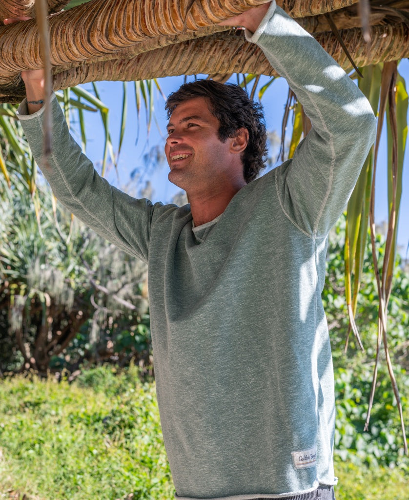 A man wearing the Golden Breed Classic Rag Top in Surf Green Marle smiles by a wooden structure outdoors, surrounded by greenery, capturing an effortless surf-inspired look.