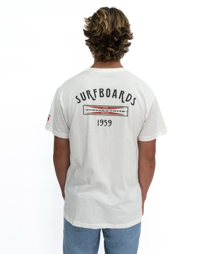 A person stands facing away, wearing the Gordon & Smith G&S Surfboards Tee in Off White with SURFBOARDS 1959 on the back, paired with light blue pants.