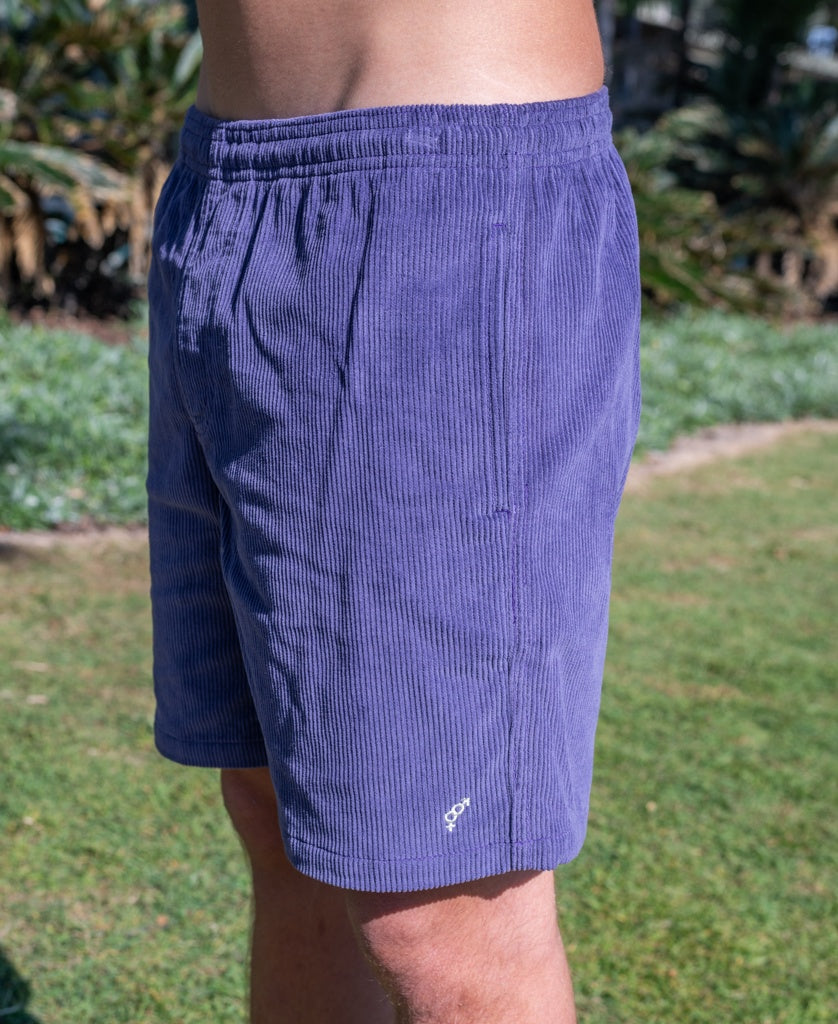A person stands outdoors in Golden Breed Floyd Cord Elastic Walkshort in Port, made from soft cotton corduroy with an elastic waistband and small white embroidered logo near the hem. Grass and plants are seen in the background.