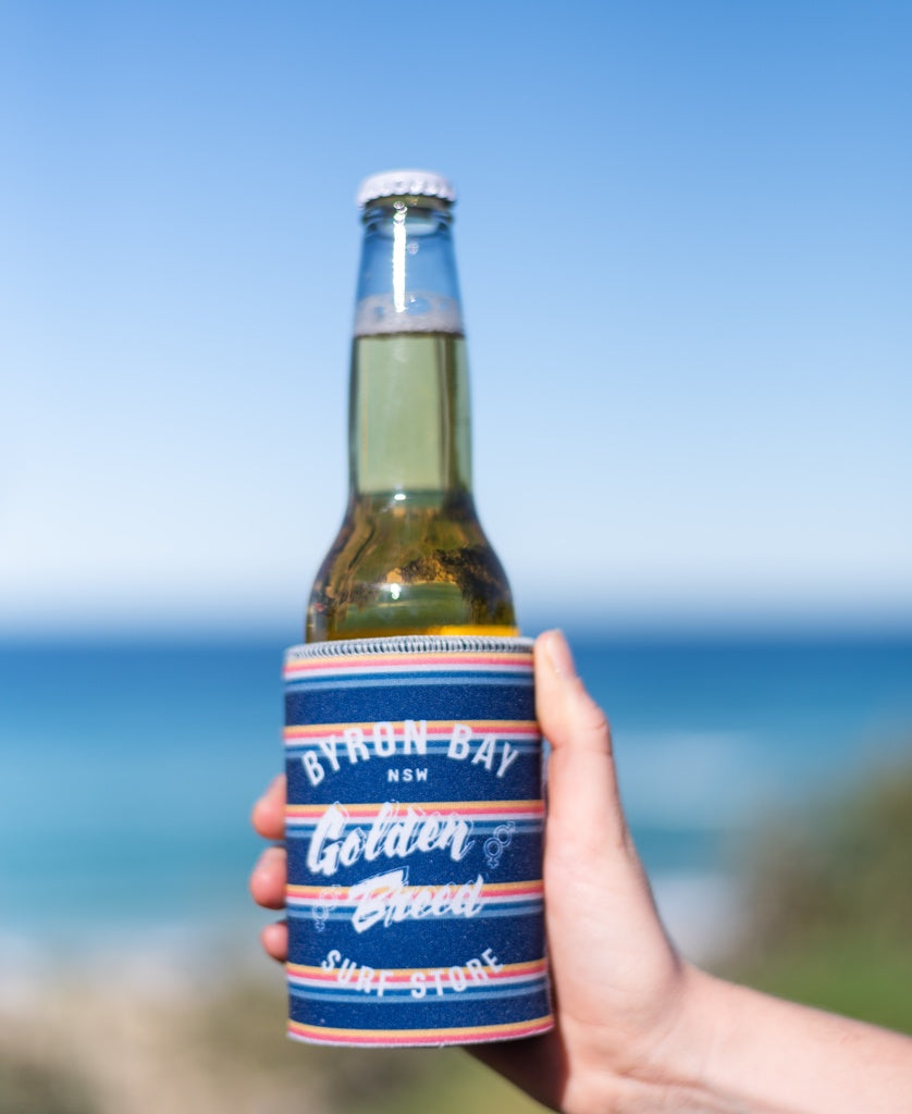 A hand holds a Golden Breed Byron Stubby Holder GB Yard in navy, wrapped around a beer bottle, set against a blurred beach and ocean background.
