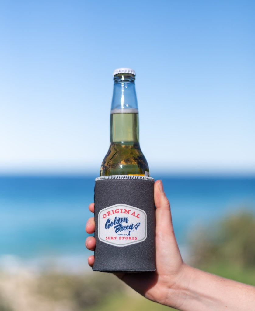 A hand holds a beer in a Golden Breed Ray Stubby Holder | Wash Black by Golden Breed, with the ocean and blue sky in the background.
