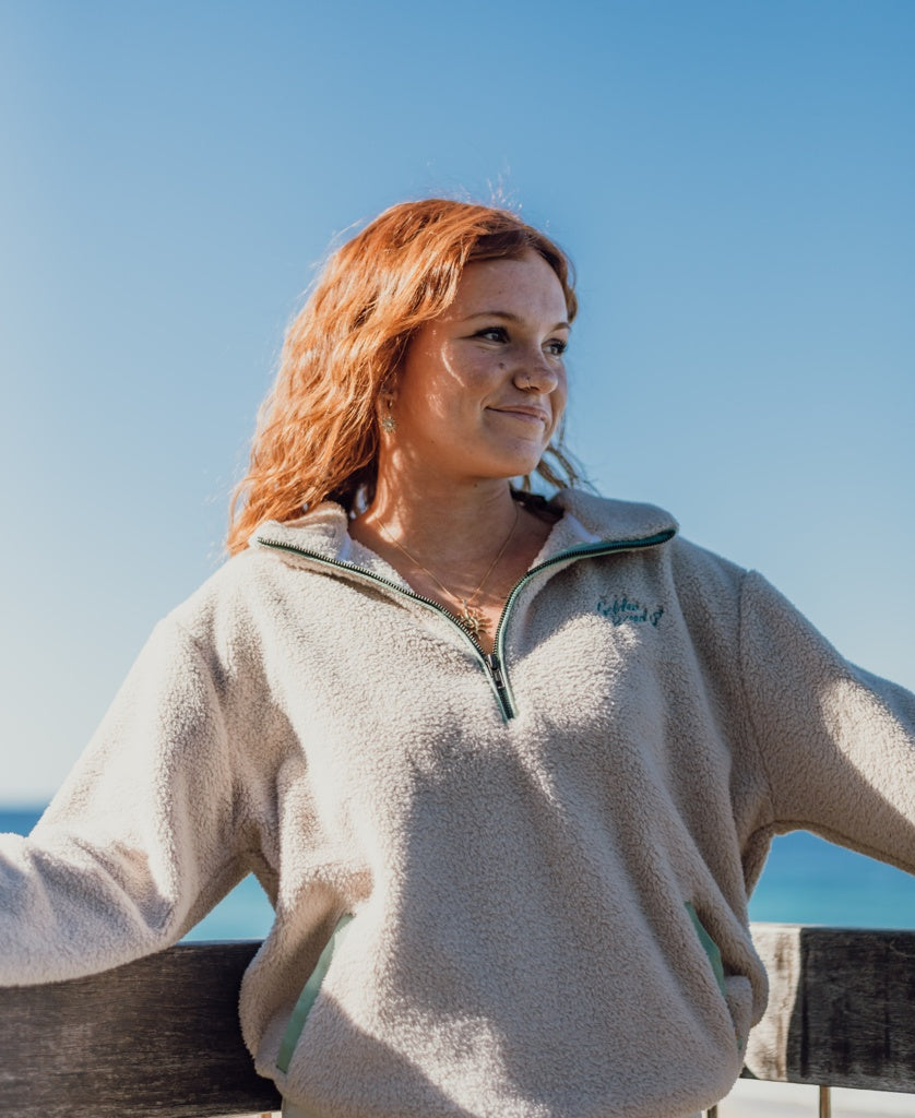 A woman with red hair wears the Golden Breed Sammie 1/2 Zip in beige, standing outdoors by a wooden railing with blue sky and water in the background.