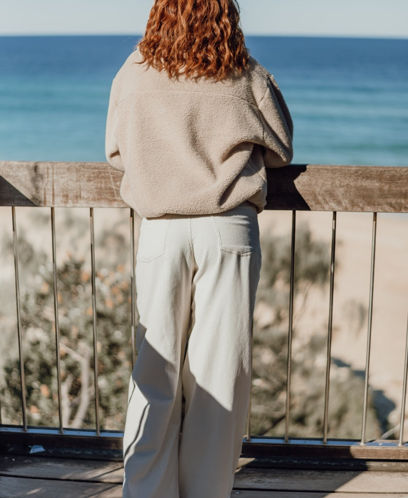A person with shoulder-length curly hair stands on a wooden deck facing the ocean, wearing the Golden Breed Sammie 1/2 Zip in beige and light-colored pants.