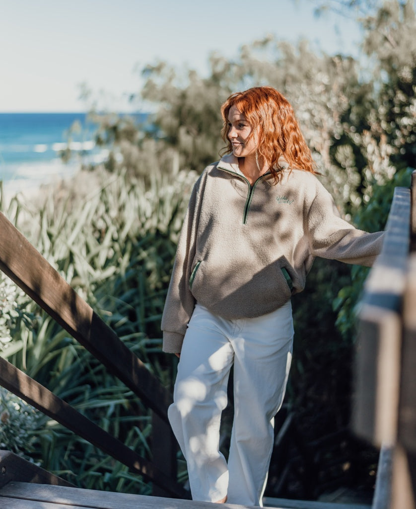 A person with red hair stands on wooden outdoor stairs near greenery, wearing the Golden Breed Sammie 1/2 Zip in Beige and white pants, with the ocean visible in the background.