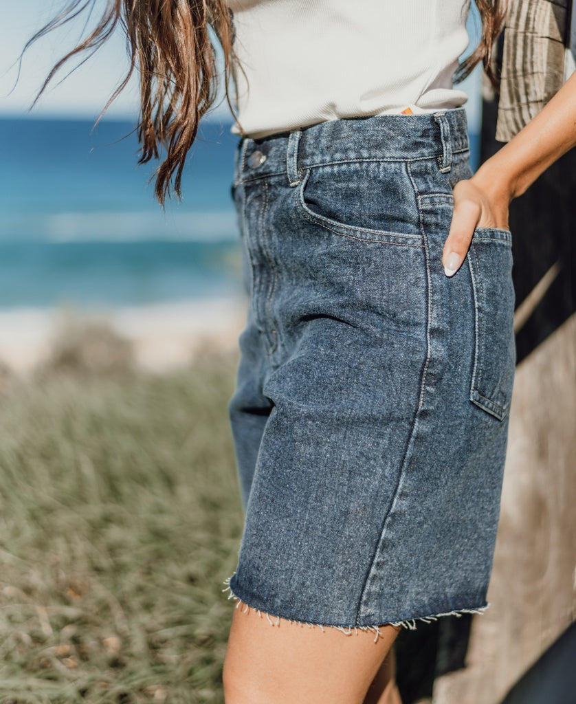 A person wearing a white top and Golden Breed Izzy Short | Dark Blue with frayed hems stands outdoors near the ocean.
