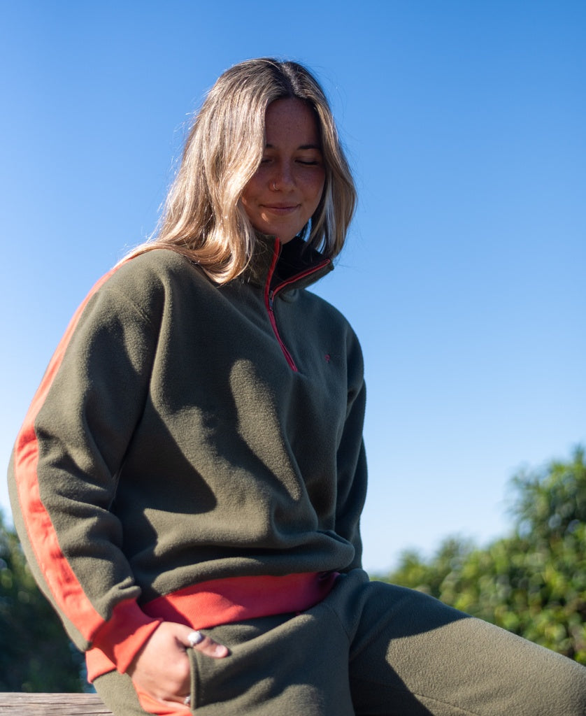 A woman in the Golden Breed Peggy 1/2 Zip Retro sweatshirt in Dk Khaki/Red sits outdoors on a sunny day, with trees and a clear blue sky behind her.