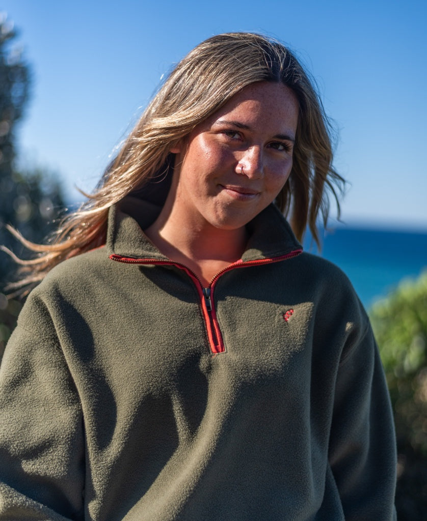 A person with long hair stands outdoors among trees and blue sky, wearing the Golden Breed Peggy 1/2 Zip Retro fleece in dark khaki with a red zipper.